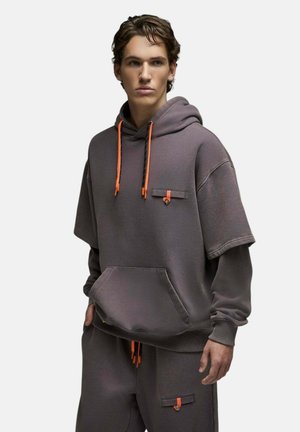Jeune homme tourné vers la gauche, portant un sweat à capuche oversize gris foncé et un pantalon assorti avec des cordons orange et des poches à boutons.