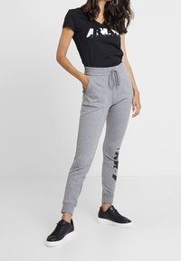 Žena v černém tričku s logem Armani Exchange, šedých šortkách se stahovací šňůrkou a černých teniskách s bílou podrážkou.