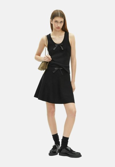 Jeune femme se tenant avec une expression neutre, portant un top sans manches noir et une jupe avec des nœuds noirs, portant un sac à bandoulière en cuir beige matelassé.