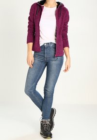 Felpa con zip in pile di colore rosa scuro con cappuccio nero, abbinata a jeans blu slim e scarponi da trekking neri con lacci grigi.