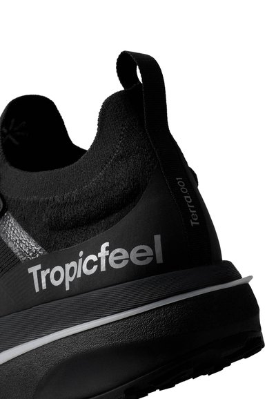Chaussure de sport noire avec une tige texturée, un logo bien visible et des accents réfléchissants ; dotée d'une languette au niveau du talon et d'une semelle rembourrée.
