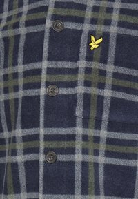 Camicia di flanella a quadri blu navy e verde con linee grigie. Presenta bottoni neri e un piccolo logo ricamato giallo. Texture morbida.