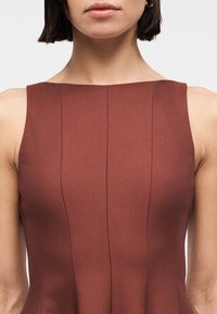 Femme portant une robe marron sans manches et ajustée, avec des détails de coutures verticales, cadrée des épaules à la taille sur un fond uni.