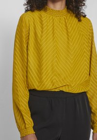 Blouse jaune moutarde avec un motif chevron texturé, col froncé et manches longues, associée à un pantalon noir à taille élastique.