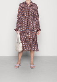 Vestido con estampado floral en navy, naranja y lavanda, con mangas largas, puños con volantes y cuello con abertura. Combinado con un bolso blanco de mimbre.