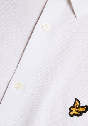Polo bianca in tessuto strutturato, con due bottoni bianchi e un logo ricamato di un uccello giallo sul petto.
