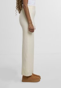 Jasno beżowe, wysokie, dzianinowe spodnie o prostym kroju i miękkiej strukturze, zestawione z brązowymi zamszowymi klapkami.