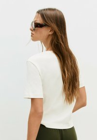 Ung kvinde med langt brunt hår iført skildpaddemønstrede solbriller, en hvid t-shirt med korte ærmer og olivengrønne bukser, vender mod venstre.