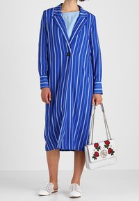 Modrý pruhovaný dlouhý blejzr s jedním černým knoflíkem, spárovaný s bílou kabelkou s červenými růžovými akcenty a bílými perforovanými slip-on botami.