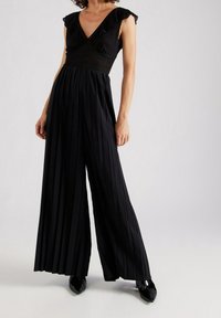 Svart jumpsuit med figursydd överdel, V-ringning, vida ben med vertikala veck och volangärmar. Glansiga svarta spetsiga skor.