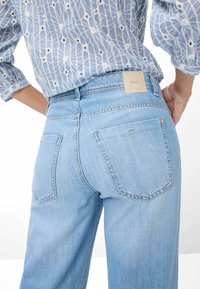 Vaqueros de denim azul claro con cintura alta, que presentan una etiqueta de cuero cuadrada, dos bolsillos traseros y detalles de costura sutiles.