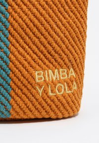 Detailný záber na oranžovú pletenú látku s diagonálnymi tyrkysovými pruhmi a žltým vyšívaným textom "BIMBA Y LOLA" v pravom dolnom rohu.