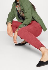 Pantaloni in corduroy rossi, giacca a scacchi verde, camicia floreale con accenti neri e mules con tacco neri. Focus sulla texture e sui colori vivaci.