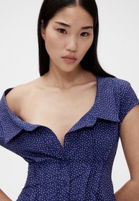 Abito blu navy a pois con bottoni sul davanti, maniche corte e colletto ampio. Presenta un corpetto aderente e spalle scoperte.