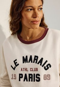 Sudadera blanca con cuello de canalé burdeos que presenta el texto "LE MARAIS ATHL CLUB PARIS 19 89" bordado en negro y burdeos.