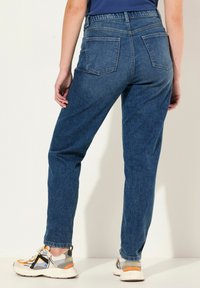 Džínové kalhoty v středně modré barvě s vysokým pasem, rovnými nohavicemi a dvěma zadními kapsami. Materiál vypadá odolně a má hladkou strukturu.