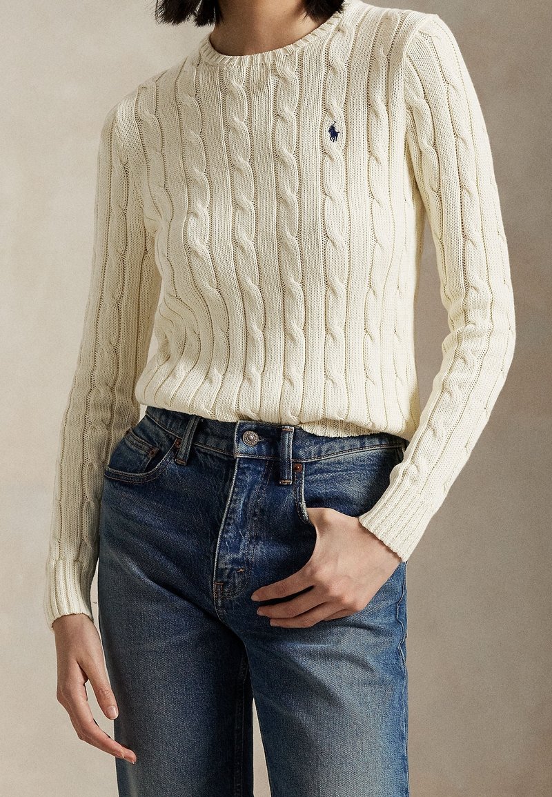 Femme portant un pull crème en maille torsadée avec un petit logo bleu marine et un jean bleu taille haute, une main dans la poche, fond neutre.