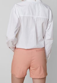 Vista trasera de una camisa blanca de manga larga, combinada con pantalones cortos rosas. Los pantalones cortos son lisos, con dos bolsillos traseros y una textura suave.
