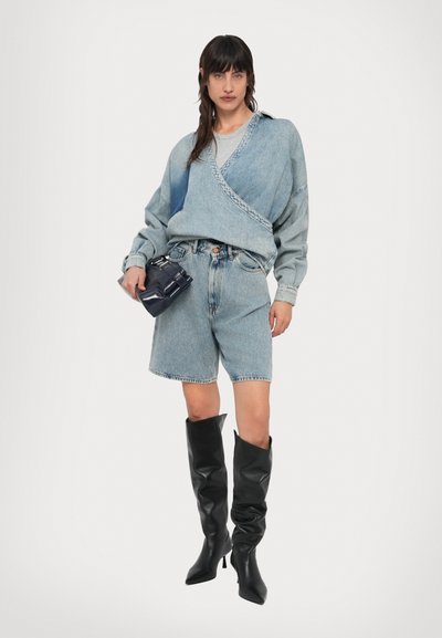 Γυναίκα με ανοιχτό γαλάζιο denim τοπ τύπου wrap και σορτς, κρατώντας σκούρο μπλε τσάντα, και φοράει μαύρες μπότες ψηλές μέχρι το γόνατο με τακούνι, στέκεται μπροστά σε λευκό φόντο.