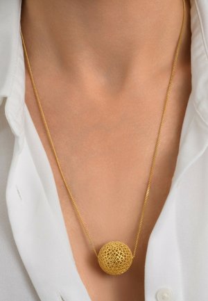 Collier en or avec un pendentif sphérique présentant un motif en treillis complexe, reposant sur un cou nu au-dessus d'une chemise blanche.
