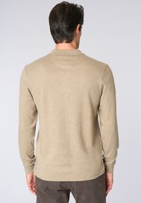 Uomo in camicia beige a maniche lunghe e pantaloni scuri, mostrato di spalle su uno sfondo grigio chiaro uniforme.