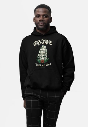 Hombre con sudadera negra con capucha con un barco de vela y el texto "Lost at Sea", combinado con pantalones negros con líneas de cuadrícula blancas.