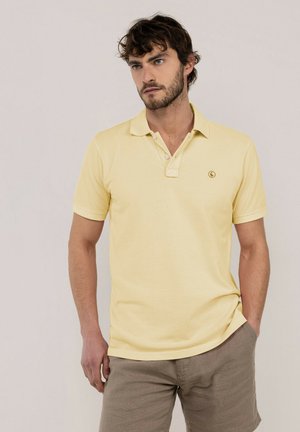 Hombre con cabello corto y oscuro y barba, que lleva una camisa polo amarilla clara y pantalones cortos beige, de pie con una mano en el bolsillo frente a un fondo liso.