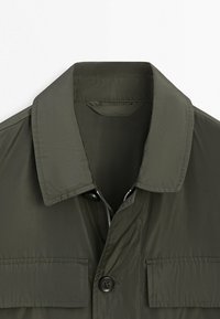 Colletto di giacca verde scuro con una cerniera parzialmente aperta e un bottone nero sotto due tasche con patta sul petto.