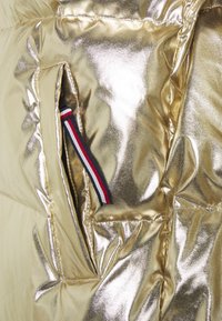 Zlatá přechodová bunda s lesklým kovovým vzhledem. Má boční kapsu odhalující detail s pruhy v červené, bílé a modré barvě.