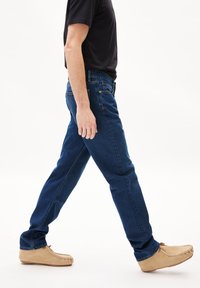 Dunkelblaue Jeans mit geradem Schnitt, getragen mit einem schwarzen T-Shirt und beigen Mokassin-Schuhen. Gehpose, Seitenansicht.
