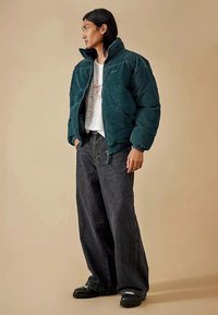 Mörkgrön pufferjacka i manchester med hög krage, dragkedja och ribbade muddar, kombinerad med vida mörka jeans och svarta skor.