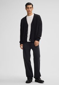 Zwarte gebreide cardigan met een V-hals, grote knopen, ribbels aan de mouwen, gedragen over een wit T-shirt en gecombineerd met zwarte broeken en laarzen.