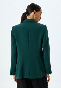 Blazer teal con una vestibilità su misura, colletto a rotondo e spalle strutturate. Presenta maniche lunghe con dettagli a bottone e schiena sfiancata.
