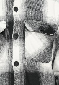 Camicia di flanella a quadri in bianco e nero con due tasche sul petto, chiusura a bottoni sul davanti e una texture morbida. Presenta bottoni neri e un motivo a scacchi.