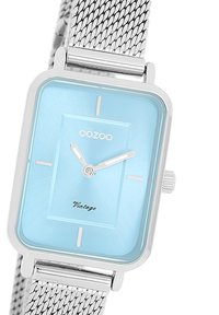 Montre carrée avec un cadran bleu clair, des index horaires en argent et un bracelet en maille métallique. Présente une surface texturée et une couronne sur le côté.