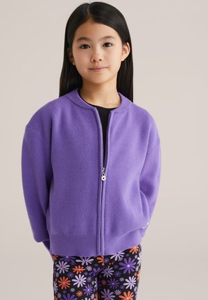 Junges Mädchen mit langen schwarzen Haaren, das einen lila Reißverschluss-Pullover und schwarze Leggings mit bunten Blumenmustern trägt, steht vor einem einfarbigen Hintergrund.