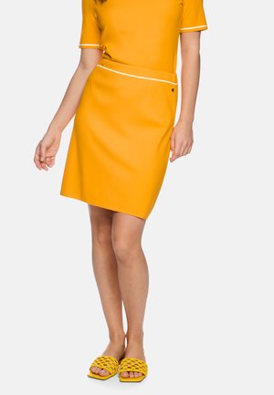 Frau trägt ein leuchtend gelbes Kurzarmoberteil, einen passenden knielangen Rock mit weißem Besatz und gelbe offene, gewebte Sandalen.