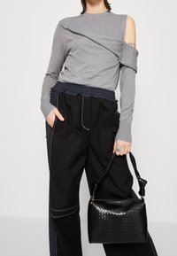 T-shirt gris à manches longues avec un design asymétrique, associé à un pantalon noir avec une taille froncée et des coutures blanches. Sac à main tressé noir.