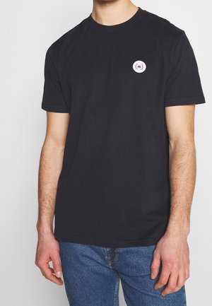 T-shirt noir en coton à manches courtes, col rond, avec un logo circulaire brodé sur le côté gauche de la poitrine. Porté avec un jean bleu.