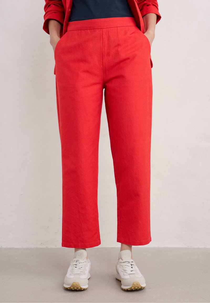 Personne portant un pantalon rouge vif et une veste assortie, les mains dans les poches, chaussée de baskets blanches sur un sol clair.