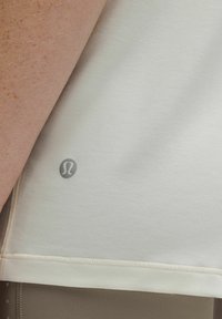 Nahaufnahme eines Arms neben einem cremefarbenen Stoff mit einem kleinen, grauen Lululemon-Logo in der Nähe der Naht.