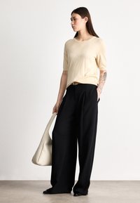 Pulover bej cu mânecă scurtă, asortat cu pantaloni negri largi, cu talie înaltă. Modelul ține o geantă mare crem tip tote. Texturi netede.