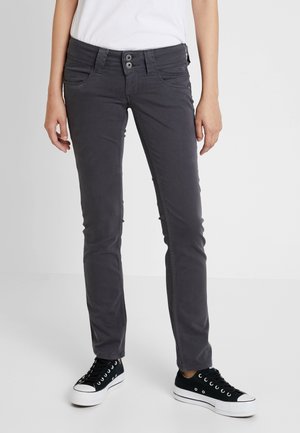 Osoba w ciemnoszarych, dopasowanych spodniach oraz czarnych, niskich trampkach z białą podeszwą, stojąca na tle jasnego, jednolitego tła.