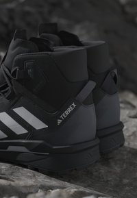 Close-up af sorte og grå Adidas Terrex vandrestøvler, der hviler på klippefyldt terræn.