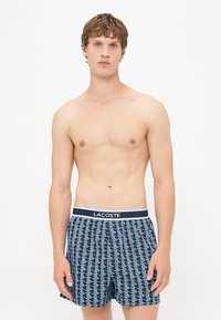 Boxeri negru marin cu un model geometric, talie albă cu logo-ul "LACOSTE", material ușor și croială lejeră.