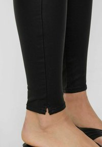 Leggings noirs avec une texture lisse, ajustement fuselé et ourlet évasé. Portés avec des sandales à lanière noires, mettant en valeur la cheville et le pied.