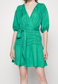 Mujer vistiendo un vestido de satén verde con mangas acampanadas hasta los codos, detalles en la cintura fruncida y un cinturón atado a la cintura.