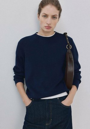 Giovane donna con capelli tirati indietro indossa un maglione blu scuro e una gonna di jeans, con le mani nelle tasche, porta una borsa a tracolla marrone scuro su sfondo grigio.