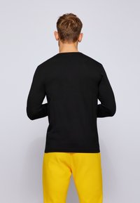 Schwarzes Langarmshirt mit Rundhalsausschnitt, kombiniert mit hellgelben Hosen. Glatte Textur, keine sichtbaren Muster oder Logos.