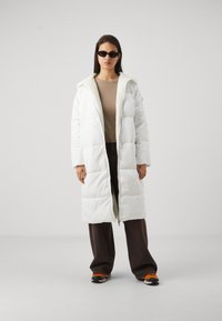 Doudoune blanche avec un col en fourrure, dotée d'une fermeture éclair, d'une coupe surdimensionnée et d'un motif matelassé. Associée à un pantalon large marron.
